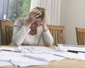 Overwhelmed woman at home stressed over paperwork and bills, sitting at a dining table with documents and crumpled papers.
