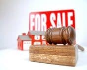 Gavel and "For Sale" sign in front of a house model, representing real estate transactions.