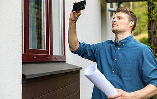 Home appraiser photographing exterior window while holding blueprints during a property inspection in Western Colorado
